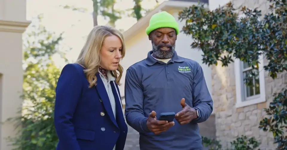 A woman in a dark blazer and a man in a green beanie are outdoors, looking intently at a phone held by the man. He wears a branded work shirt as they review the screen together.