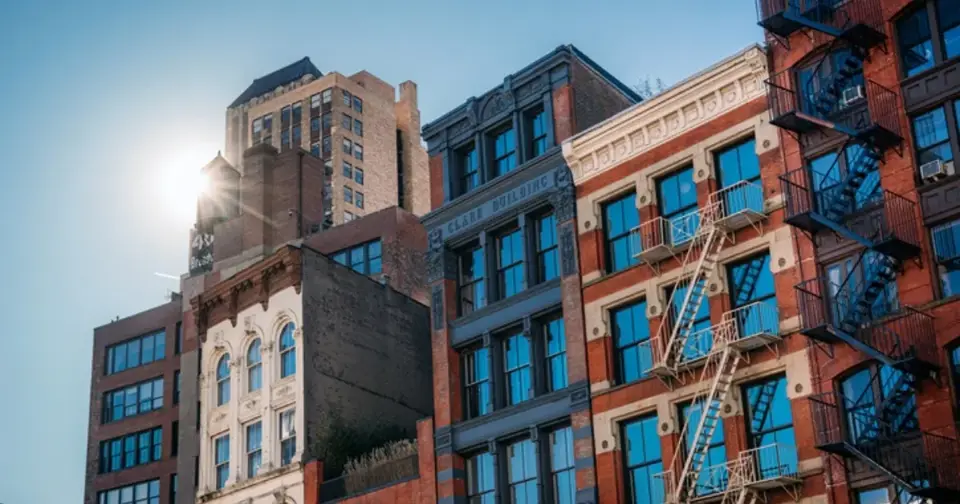 Upper parts of several urban apartment buildings with bright sunlight. The buildings feature brick and stone facades, numerous windows, and a visible fire escape.